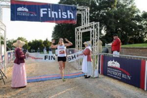 A woman crosses the finish line of the George Washington Patriot Run breaking a ribbon held by two women dressed in period costume. (Photo: Mount Vernon)