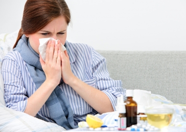 A Sick woman in bed blowing her nose surrounded by a lemon, pills, nose spray and cough medicine. (Photo: Getty Images)