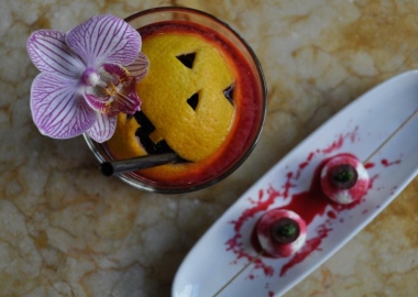 Cocktail with a jack-o-lantern face carved into an orange peel floating in itand two vampire bites pintxos that look like bloody skewered eyeballs on a plate beside it. (Photo: Estadio)