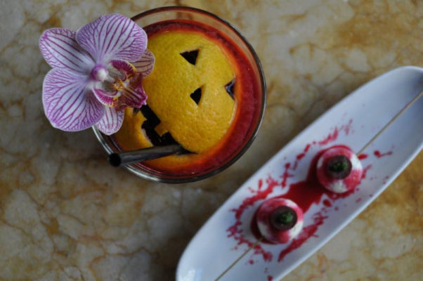 Cocktail with a jack-o-lantern face carved into an orange peel floating in itand two vampire bites pintxos that look like bloody skewered eyeballs on a plate beside it. (Photo: Estadio)