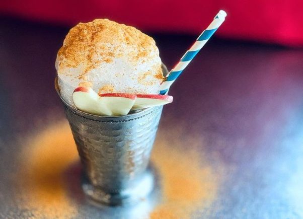 A drink in a metal cup topped with foam, baking spices and apple slices with a blue and white straw. (Photo: The Royal)