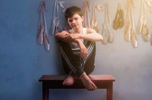 Billy Elliot sitting cross-legged on a table with ballet slippers hanging on the wall behind him. (Photo: Signature Theatre)