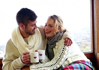 man and woman in white sweaters and scarves cudding in a ski lodge with cups of coffee and a blanket. (Photo: Deposit Photo)