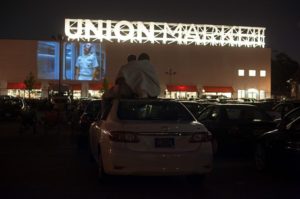 Two people sitting on the roof of a car wrapped in a blanket watching a movie projected on the outside of Union Market. (Photo: Union Market)