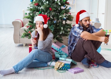 Unhappy woman and man sitting back-to-back infront of a Christmas tree wearing Santa hats and with cocktails. (Photo: Shutterstock)