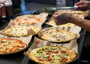 An employee sprinkles pesto on top of a cooked pizza with 5 other cooked pizzas on the counter. (Photo: MOD PIzza)
