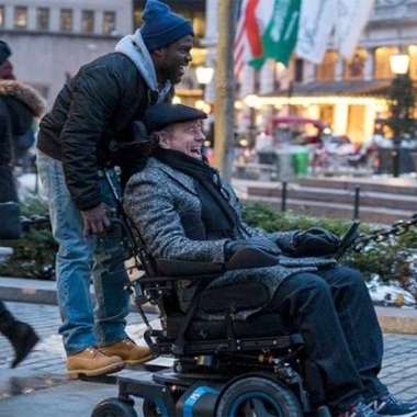 Kevin Hart standing on the back of a wheelchir driven by Bryan Cranston in The Upside. (Photo: The Weinstein Co.)
