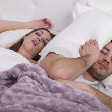 A couple in bed. The woman is snoring and the man has a pillow wrapped around his head. (Photo: iStock)