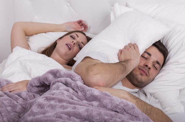 A couple in bed. The woman is snoring and the man has a pillow wrapped around his head. (Photo: iStock)