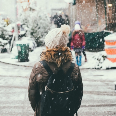 Woman in a brown winter jacket standing at the cornery of a snow-covered road. (Photo: Joseph Pearson/Unsplash)