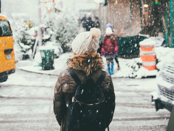 Woman in a brown winter jacket standing at the cornery of a snow-covered road. (Photo: Joseph Pearson/Unsplash)