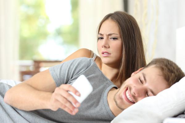 Man lying in bed toward camera texting on smart phone while woman behind him peers over shoulder. (Photo: Shutterstock)