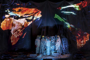 Performers in costume on stage with a map of the continents projected around them. (Photo: Johan Persson)