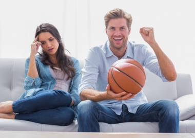 A man and woman watching a basketball game on TV. She looks disinterested while he is cheering and holding a basketball. (Photo: Shutterstock)
