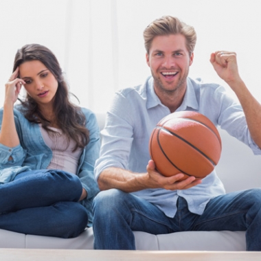 A man and woman watching a basketball game on TV. She looks disinterested while he is cheering and holding a basketball. (Photo: Shutterstock)