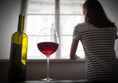 A woman stares out a window. Behind her, in the foreground, a glass of wine sits next to a bottle. (Photo: Getty Images)