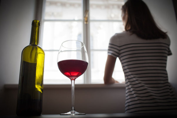 A woman stares out a window. Behind her, in the foreground, a glass of wine sits next to a bottle. (Photo: Getty Images)