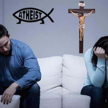 A sad couple sitting on a couch with an athesist symbol behind him and a cross behind her. (Photo: Shutterstock and Patheos)