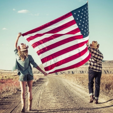 Young couple wearing cowboy hats carry a huge US flag down a dirt road towards some plateaus. (Photo: Getty Images)