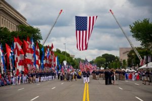Parade participants pass under a huge American flag suspended over the street by two cranes. (Photo: American Veterans Center)