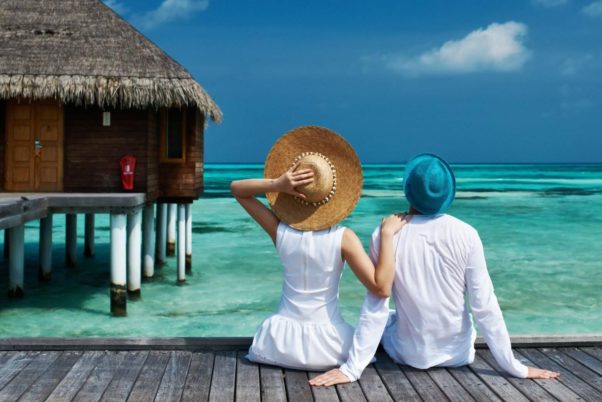 Couple dressed in white wearing hats sitting on a dock in front of a thatch hut looking out at clear blue water & a blue sky. (Photo: Getty Images)