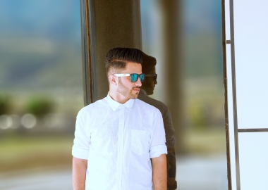 A man leaning against a glass wall with a white Oxford shirt with the arms rolled up and sunglasses. (Photo: Awar Jahfar/Unsplash)