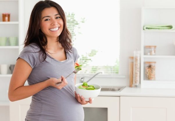 A pregnant stnding in the kitchen eating a salad. (Photo: Shutterstock)