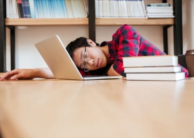 An Asian boy sleeping in the library with his laptop open on a table beside a pile of books. (Photo: Footage Firm Inc.)