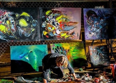 A man paining a picture with several pictures diplayed behind him. (Photo: Art All Night)