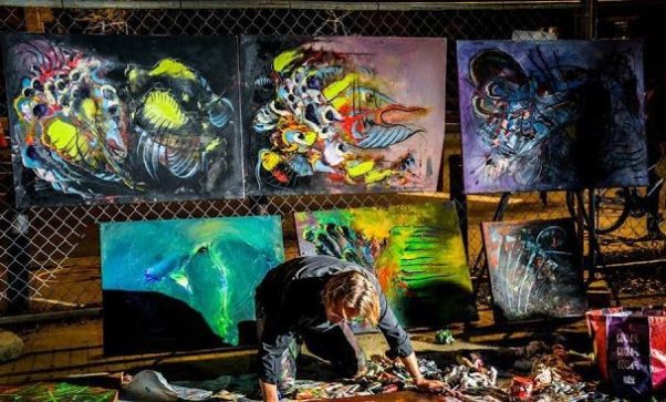 A man paining a picture with several pictures diplayed behind him. (Photo: Art All Night)