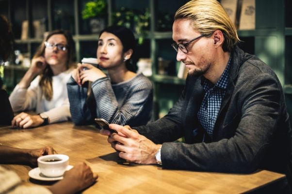 Blond man with glasses at a table texting with two women sitting beside him. (Photo: rawpixel/Pexels)