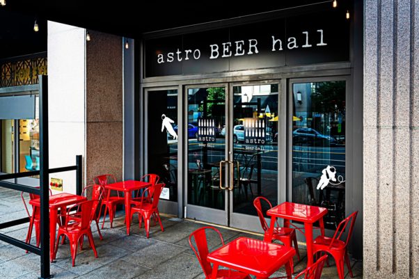 Enterance to Astro Beer Hallo with name above doors glass doors and red metal tables and chairs outside. (Photo: Scott Suchman)