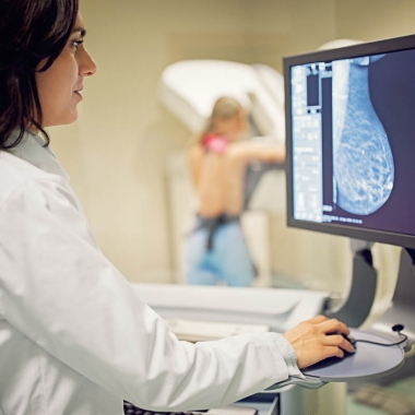 Doctor looking at a monitor while a woman in the background gets a mammogram. (Photo; Getty Images)