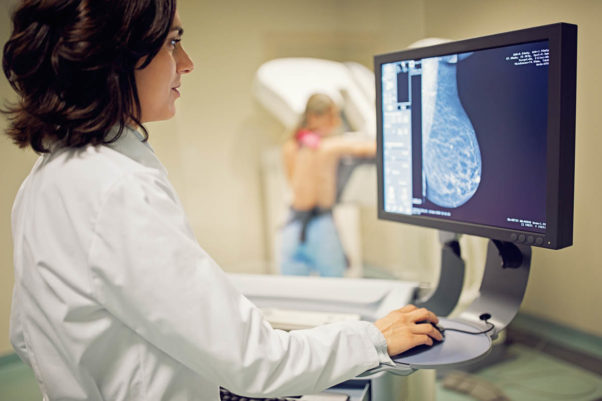 Doctor looking at a monitor while a woman in the background gets a mammogram. (Photo; Getty Images)