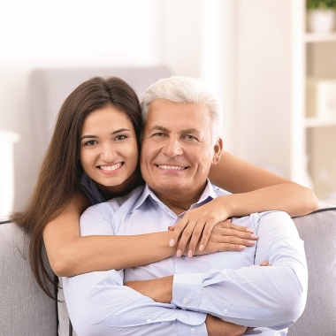 Younger woman with arms around older man who is sitting on a couch. (Photo: Shutterstock)