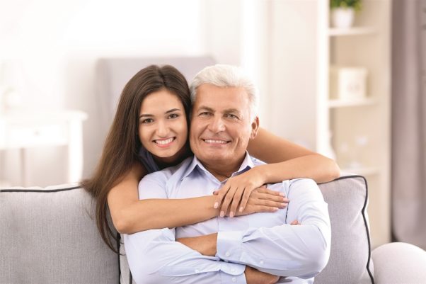 Younger woman with arms around older man who is sitting on a couch. (Photo: Shutterstock)
