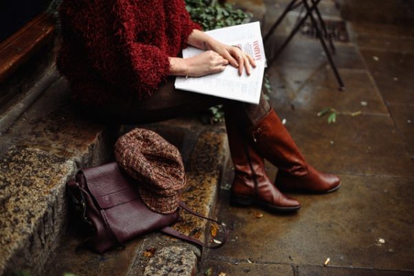 A woman sitting on steps in a sweat, leggins and thigh-high boots reading a magazine with her brief case and hat beside her. (Photo: Daria Shevtsova/Pexels)
