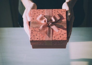 Two hands holding out a present wrapped in dark red paper and a bow. (Photo: Porapak Apichodilok/Pexels)