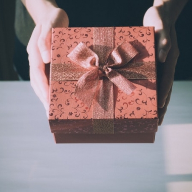 Two hands holding out a present wrapped in dark red paper and a bow. (Photo: Porapak Apichodilok/Pexels)