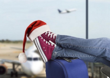 Man's feet in red Converse sneakers propped on a blue suitcase with a Santa hat over the toes and airplanes on the runway out through the window. (Photo: Shutterstock)