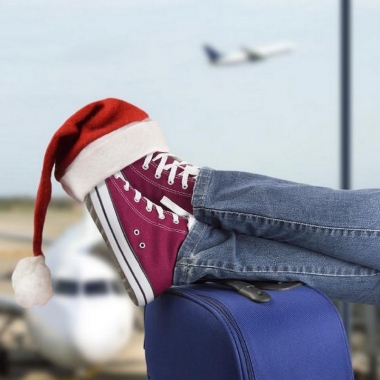 Man's feet in red Converse sneakers propped on a blue suitcase with a Santa hat over the toes and airplanes on the runway out through the window. (Photo: Shutterstock)