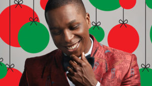 Photo of Leslie Odom Jr. from the shoulders up with his chin on his hand in from of a background of red and green ornaments. (Photo: Kennedy Center)