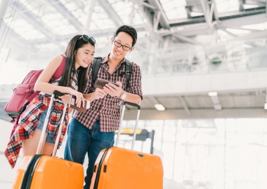 Asian couple in airport checking their cell phone while standing with suitcases. (Photo: Sushiman/Shutterstock)