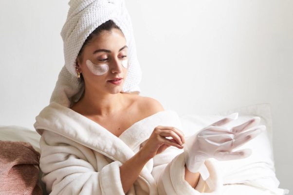 Woman in a white robe with towel wrapped around her head applying hand masks. (Photo: skincare.com)