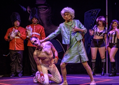 A man dresses as Queen Elizabeth in dress and pearls punches a man on his knees dressed like Freddy Mercury while female referees in shorts and halter tops and palace guards watch. (Photo: AstroPop)