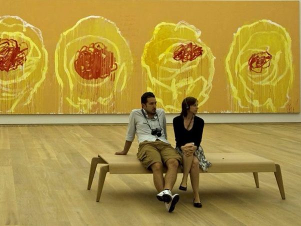 A bored man with a camera around his neck sits next to a woman on a bench who is listening to audio about a painting. (Photo: Digital Cat/Flickr)