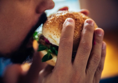 A close-up of a man with a short beard and mustache holding a burger on a bun, about to take a bite out of it. He holds the burger with both hands. (Photo: Getty Images)