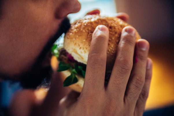 A close-up of a man with a short beard and mustache holding a burger on a bun, about to take a bite out of it. He holds the burger with both hands. (Photo: Getty Images)