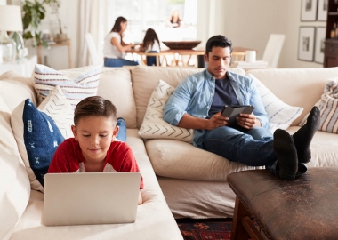 Man sitting on couch with feet propped on table reading tablet while boy lies on stomach using laptop with woman sitting at table in background. (Photo: Getty Images)