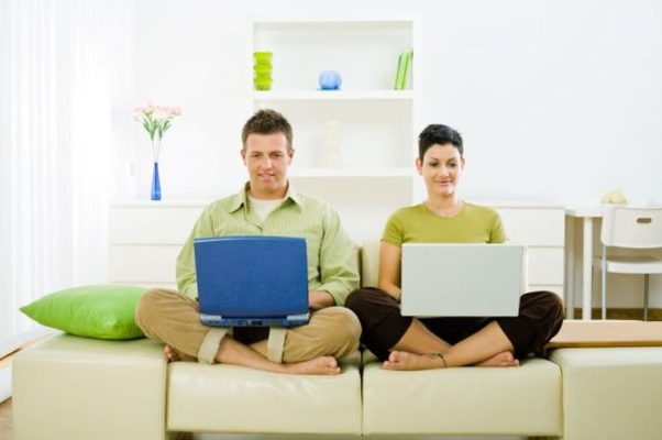 Man and woman working side-by-side on laptops while sitting on a white couch. (Photo: Shutterstock)
